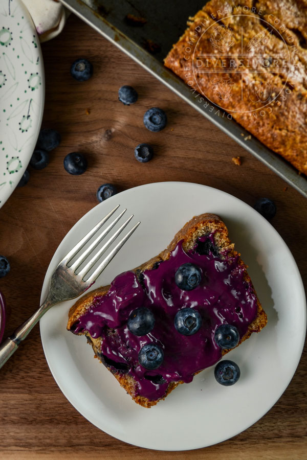 A slice of blueberry juniper rye cake on a white plate with an old fork, topped with blueberry-lemon curd and fresh blueberries